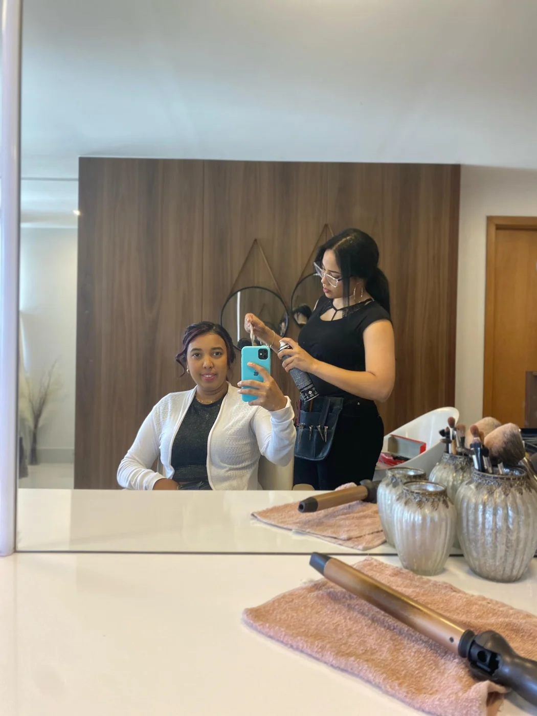 Estúdio refletido no espelho: uma cliente sentada e a maquiadora finalizando o penteado com spray, diante da parede de madeira com espelhos redondos