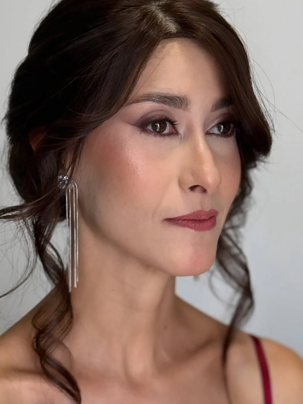 Cliente com olhos marcados em tons de ameixa, batom vinho e brincos longos, cabelo preso com mechas soltas