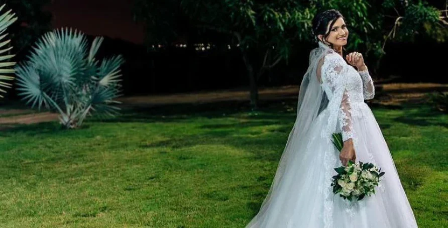Noiva de vestido branco e véu de renda segurando o buquê num jardim
