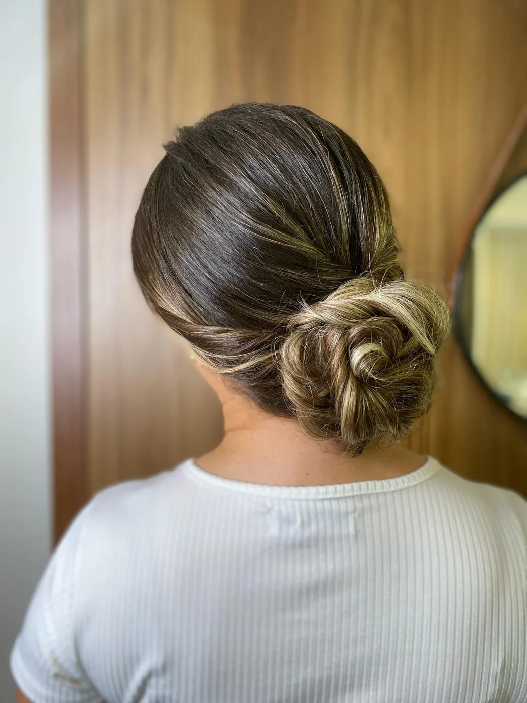 Coque baixo torcido em cabelo com mechas iluminadas, visto de costas diante da porta de madeira