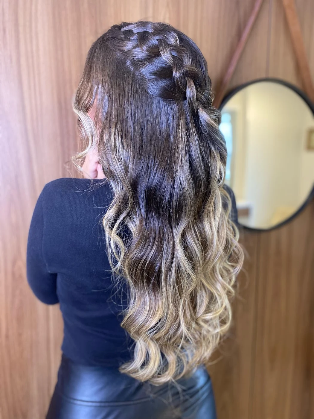 Trança no alto da cabeça e ondas longas com mechas claras, vista de costas diante do espelho redondo